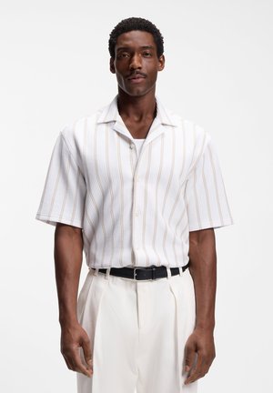Άνδρας που φοράει λευκό πουκάμισο με κοντό μανίκι, κουμπωτό, με λεπτές μπεζ ρίγες και λευκό πλυμένο παντελόνι με μαύρη ζώνη.