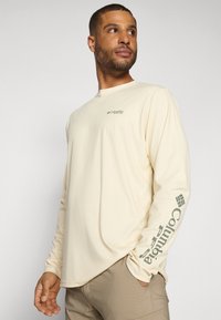 Camicia a maniche lunghe beige chiaro con scollo rotondo e logo verde Columbia stampato sulla manica destra. Tessuto morbido e leggero.