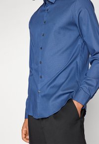 Camicia azzurra con bottoni, in tessuto strutturato, dal design aderente, con taschino sul petto e bottoni neri. Maniche lunghe con polsini a bottoni.