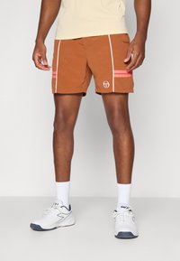 Mænds brune badebukser med hvide side-striber, lyserøde detaljer og logo. Stoffet ser let og hurtigttørrende ud. Båret med hvide træningssko.