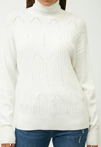 White cotton-blend knit sweater with a high ribbed collar, textured cable patterns, and a relaxed fit, paired with dark denim jeans.