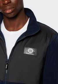 Veste zippée marine présentant une combinaison de tissu lisse et texturé, avec un patch logo sur la poitrine et un col montant.