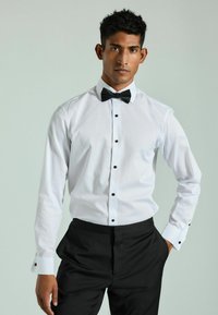 Cămașă albă cu butoni negri, asortată cu pantaloni negri și o papion negru. Țesătură fină, design structurat, aspect formal clasic.