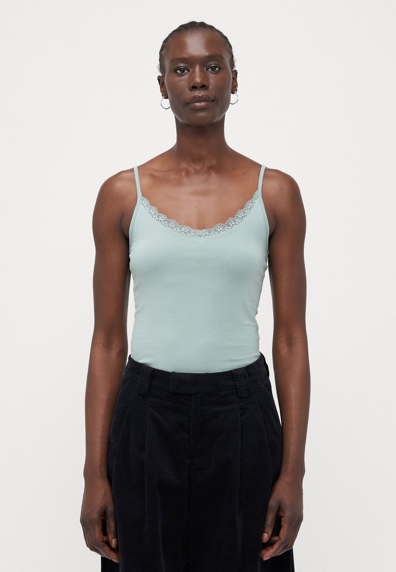 Lys blå tanktop med en scalloped blonde trim ved halsudskæringen, med tynde stropper. Sat sammen med sorte højtaljede bukser.