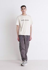 Kremfarget t-skjorte med "PEPE JEANS" trykk, kombinert med grå cargo-bukser og grå sneakers. Glatt stoff, løs passform og funksjonelle lommer.