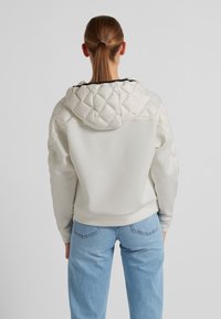 Gepolsterter cremefarbener Kapuzenpullover mit glattem Körper, strukturiertem Kapuze und elastischen Bündchen, kombiniert mit hellblauen Jeans.