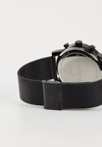Bracelet de montre en maille en acier inoxydable noir avec un système de fermeture. L'arrière présente une surface réfléchissante argentée avec une gravure de la marque.