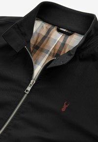 Veste noire zippée avec doublure intérieure à carreaux marron et petit logo cerf rouge sur la poitrine près de la fermeture éclair et du col.