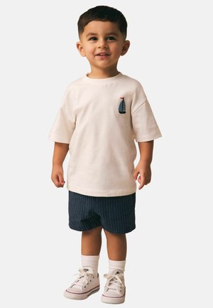 Junger Junge steht, trägt ein weißes T-Shirt mit einem kleinen Segelboot-Emblem, marineblaue gestreifte Shorts, weiße Socken und weiße Turnschuhe.