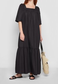 Robe maxi noire avec un décolleté carré, des manches bouffantes et un ourlet à volants. Tissu doux, coupe décontractée, associée à un sac en osier et des sandales plates.