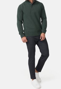 Grüner Strickpullover mit Henley-Kragen und drei Knöpfen, kombiniert mit dunkelgrauen Slim-Fit-Hosen und weißen Sneakern.