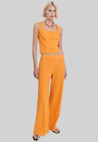 Oranje cropped tanktop met vierkante halslijn en knopen, gecombineerd met wijde broek. Gladde textuur, minimalistisch ontwerp.