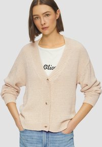 Cardigan en tricot rose pâle avec un col en V, bouton sur le devant et des poignets côtelés. Tissu texturé, coupe décontractée, porté sur une chemise blanche avec un texte noir.