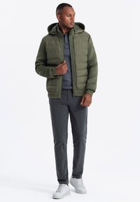 Grön puffjacka med huva, strukturerad tyg, framzipper, lager på en grå tröja, kombinerad med grå byxor och vita sneakers.