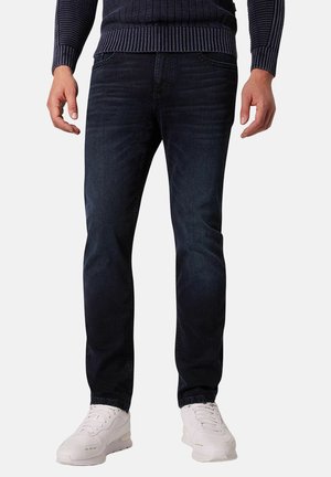 Mann trägt dunkelblaue Jeans, weiße Sneakers und einen marineblauen, gerippten Pullover, steht vor einem einfarbigen Hintergrund.