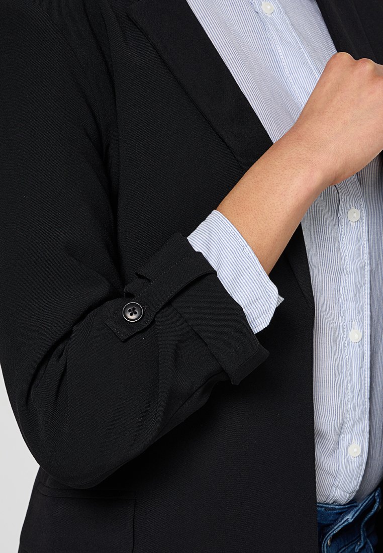 Personne portant un blazer noir avec les manches retroussées sur une chemise boutonnée à rayures bleu clair, la main près de la poitrine.