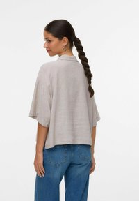 Chemise en lin gris clair avec une coupe décontractée, manches courtes et une texture subtile, associée à un pantalon en denim bleu. Aucun motif visible.