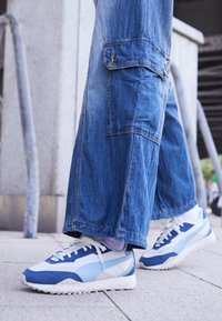 Adidași alb-albaștri cu talpă texturată. Au un design cu panouri și șireturi în față, asortați cu pantaloni cargo albaștri din denim, largi.