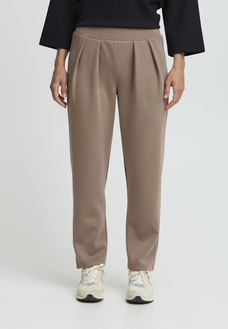 Pantaloni marroni a vita alta realizzati in materiale morbido, con design plissettato sul davanti, gambe taperate e vita elastica, abbinati a sneakers chiare.