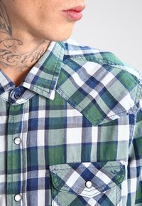 Camisa a cuadros verde y azul marino hecha de tejido tejido; presenta un cuello abotonado, botones metálicos y dos bolsillos en el pecho con solapas.