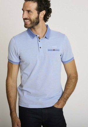 Homme aux cheveux bouclés et à la barbe, portant un polo bleu clair avec un col et des bordures de manches bleu foncé, souriant et regardant vers sa gauche.