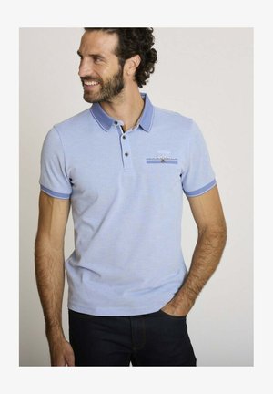 Homme aux cheveux bouclés et à la barbe, portant un polo bleu clair avec un col et des bordures de manches bleu foncé, souriant et regardant vers sa gauche.