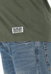 Olivgrön T-shirt med en vävd etikett som säger "DENIM FACTORY," ihop med blå denimjeans med en subtil textur och sömmar.