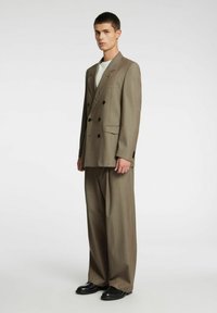Brązowy dwurzędowy garnitur z dużymi klapami, przednimi kieszeniami i czarnymi guzikami. Luźne spodnie o gładkiej fakturze. Czarna obuwie.