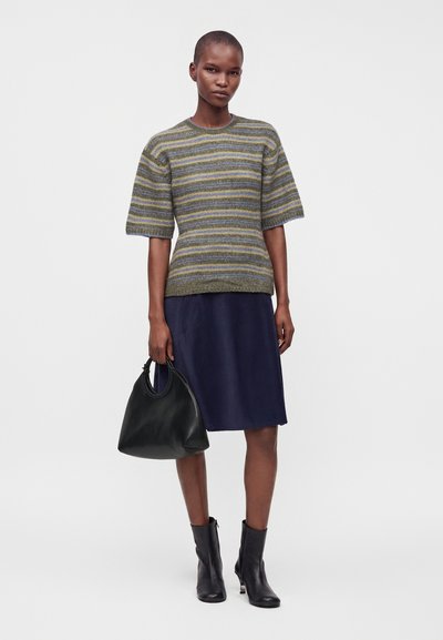 Femme portant un pull rayé à manches courtes, une jupe bleu marine mi-longue, des bottines noires et tenant un sac à main noir sur un fond uni.