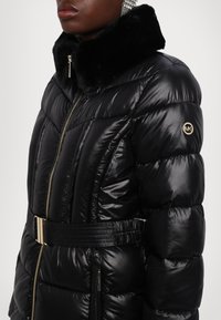 Personne portant une doudoune noire brillante avec un col en fourrure, une fermeture éclair dorée, une ceinture avec boucle et un logo MK rond sur la manche.