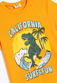 Camiseta de algodón naranja con un gráfico de un dinosaurio verde surfeando una ola, con el texto "CALIFORNIA SURF & FUN" y palmeras de fondo.