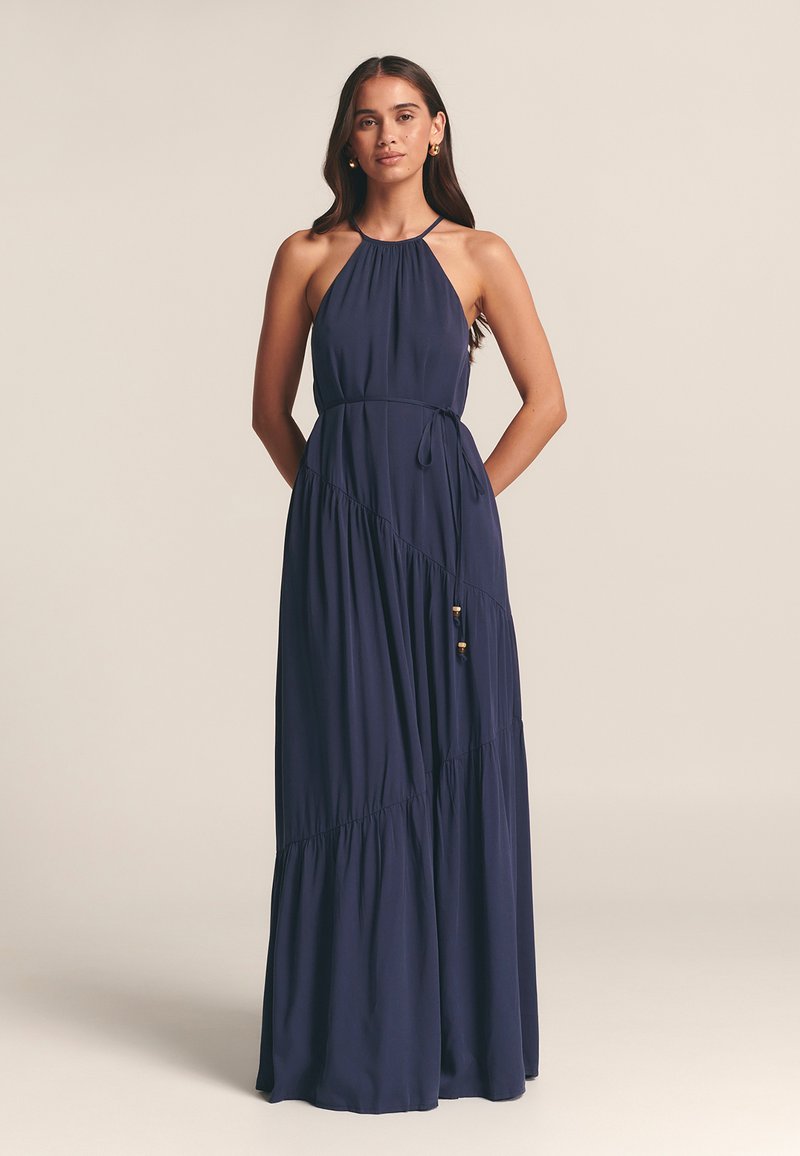 Marinblå halterneck-maxiklänning med en flödande kjol, markerad midja och dekorativa dragskoaccents. Mjukt tyg med lager-på-lager designinslag.