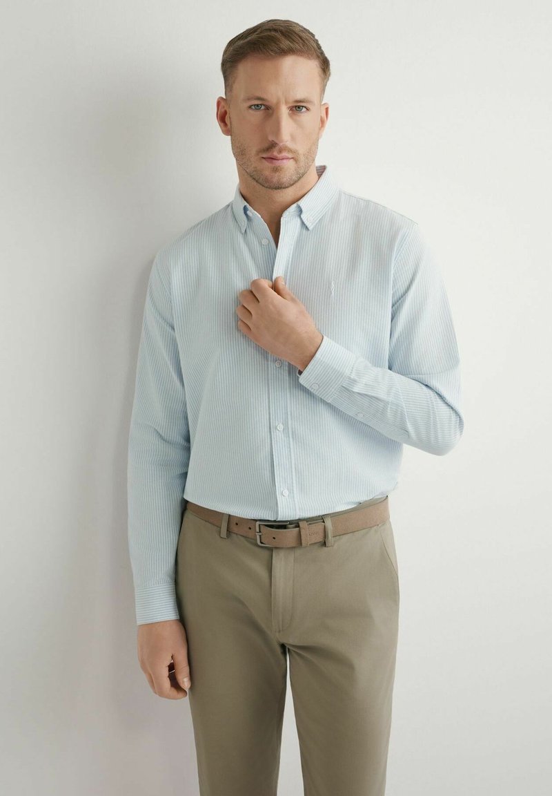 Mężczyzna poprawiający kołnierz jasnoniebieskiej koszuli w paski, ubrany w spodnie khaki i brązowy pasek, stoi na białym tle.