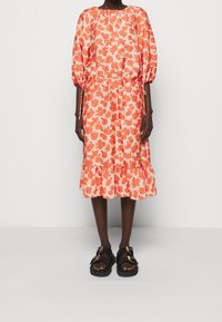 Kukkanen mekko, jossa on oransseja kukkia vaalealla taustalla, kankea vyötärö, puffihihat ja röyhelöhelma; yhdistettynä mustiin sandaaleihin.