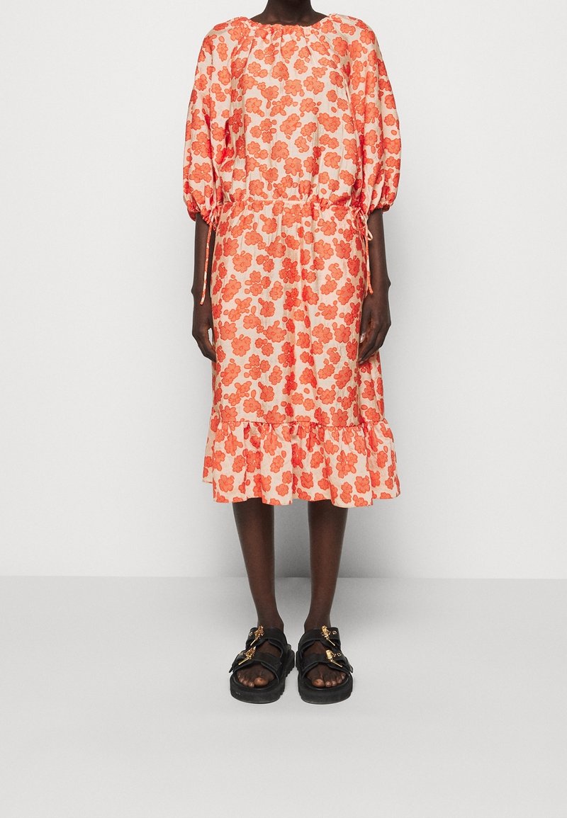 Kukkanen mekko, jossa on oransseja kukkia vaalealla taustalla, kankea vyötärö, puffihihat ja röyhelöhelma; yhdistettynä mustiin sandaaleihin.