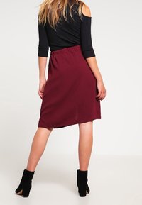 Jupe mi-longue bordeaux avec taille élastique, en tissu léger, et coupe droite. Portée avec un haut noir à épaules dénudées et des bottines.