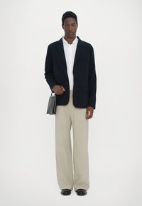 Navy blazer over a white shirt, paired with light beige wide-leg trousers. The subject holds a small black bag. Smooth textures, tailored design.