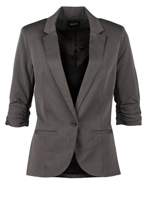 Kvinde iført en grå blazer, sort top, blå skinny jeans og sorte ankelstøvler, der holder en sort clutch i den ene hånd.