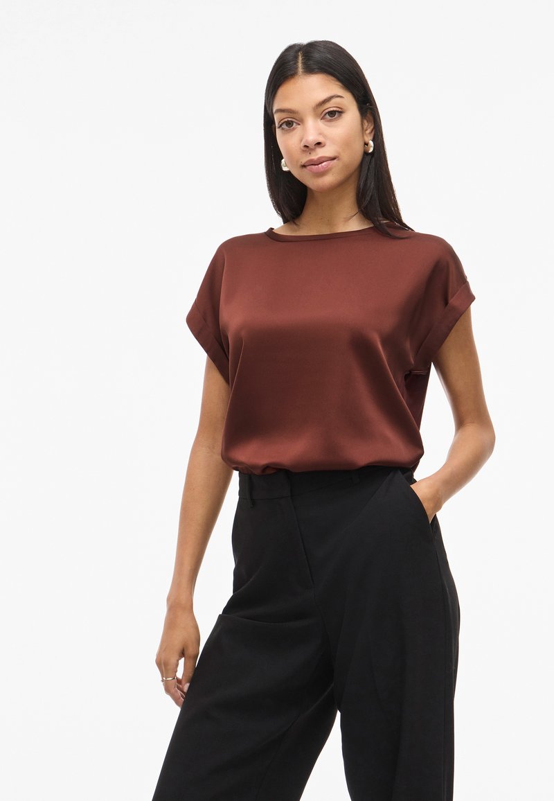 Top marrone in raso con maniche corte, scollo rotondo e polsini arrotolati, abbinato a pantaloni neri a vita alta con tasche laterali.