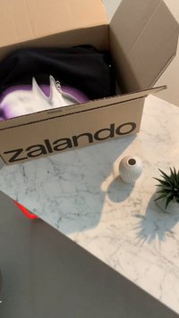 Cardboard box with "Zalando" logo, partially open revealing a black garment. White ribbed vase and small plant on a marble tabletop.