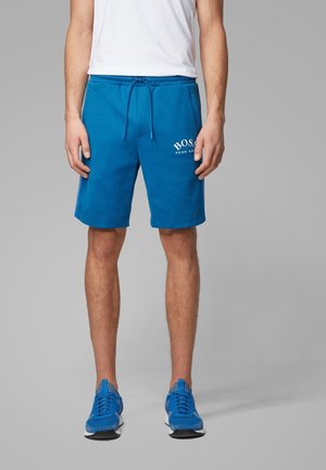 Mand iført blå Hugo Boss-shorts, hvid t-shirt og blå sneakers, stående mod en ensfarvet grå baggrund.