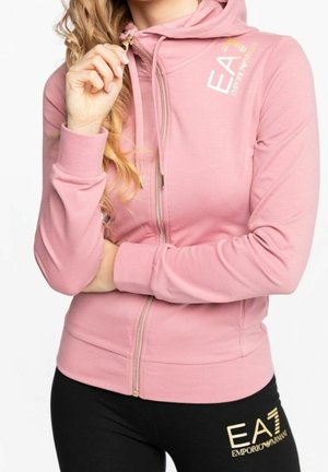 Kobieta ubrana w różową bluzę z kapturem zapinaną na zamek oraz czarne legginsy, oba oznaczone logo EA7 Emporio Armani w złotym kolorze.