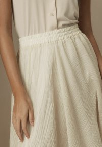 Femme portant un haut sans manches crème rentré dans une jupe crème taille haute et texturée avec une taille élastique.