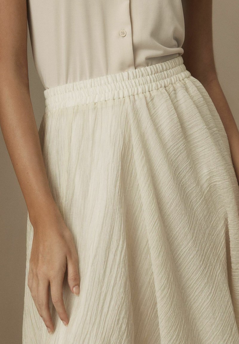 Femme portant un haut sans manches crème rentré dans une jupe crème taille haute et texturée avec une taille élastique.