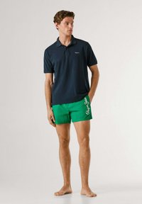 Noor mees seisab paljajalu, seljas tumesinine polosärk ja eredrohelised ujumispüksid valge logoga külje peal.