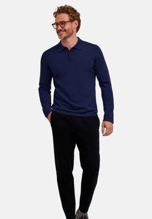 Man with glasses wearing a navy long-sleeve polo shirt and black pants standing with one hand in pocket, smiling and looking to the side.