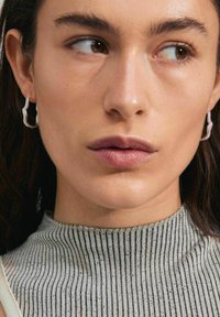 Pendientes de aro plateados con un diseño ondulado y acentos brillantes, sobre un fondo de una blusa gris texturizada y a rayas.
