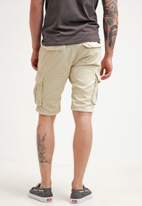 Shorts cargo beiges en coton, avec des poches latérales, une coupe décontractée et des ourlets retroussés. Portés avec des chaussures en toile grises.