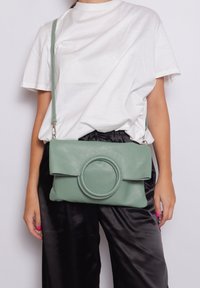 Borsa a tracolla in pelle verde con un accento circolare, forma rettangolare e chiusura a pattina. Materiale testurizzato, tracolla regolabile, design minimalista.
