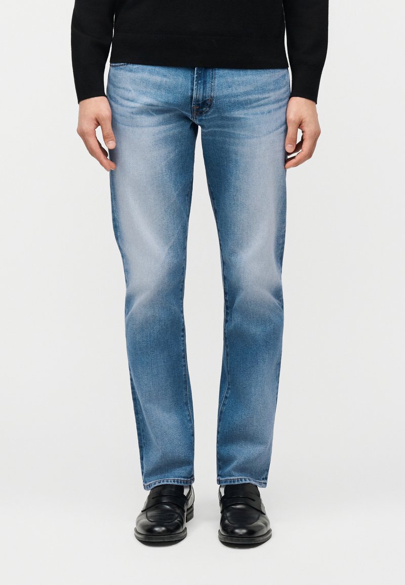 Άνδρας που φοράει ανοιχτό μπλε ξεθωριασμένο τζιν, μαύρο πουλόβερ και μαύρα δερμάτινα παπούτσια, στέκεται μπροστά σε απλό λευκό φόντο.